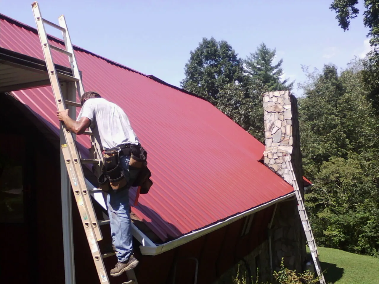 Roofer on a ladder beside a red metal roof and stone chimney — Soffit & Fascia Repair by Roofing Brothers in Zenda